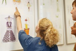 Schülerin zeigt auf eine Modezeichnung an der Ausstellungswand – gemeinsam mit Mitschüler:innen bei der Vernissage.