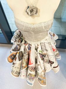 Detailaufnahme eines Kleides aus Zeitungspapier mit gefalteten Papierrosen – nachhaltiges Modekunstwerk in der Ausstellung.