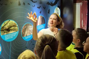 Vor einer Fledermaus-Infotafel erklärt eine Frau einer Gruppe Kinder etwas.