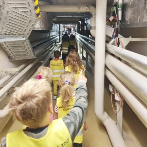 Kinder laufen durch einen Kellergang, in dem Leitungen und Rohren entlangführen.