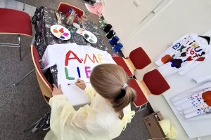 Ein Mädchen schreibt bunte Buchstaben mit einem Pinsel auf ein weißes T-Shirt.