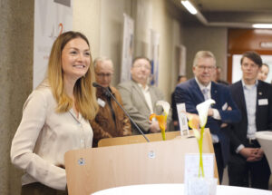 Judith Gerlach, Bayerische Staatsministerin am Mikrofon. Weitere Veranstaltungsgäste sind unscharf im Hintergrund zu sehen.