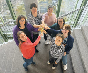 Die Praxisanleiterinnen stehen im Kreis auf ein einer Treppe und stoßen lachend mit Sektgläsern in der Mitte an.